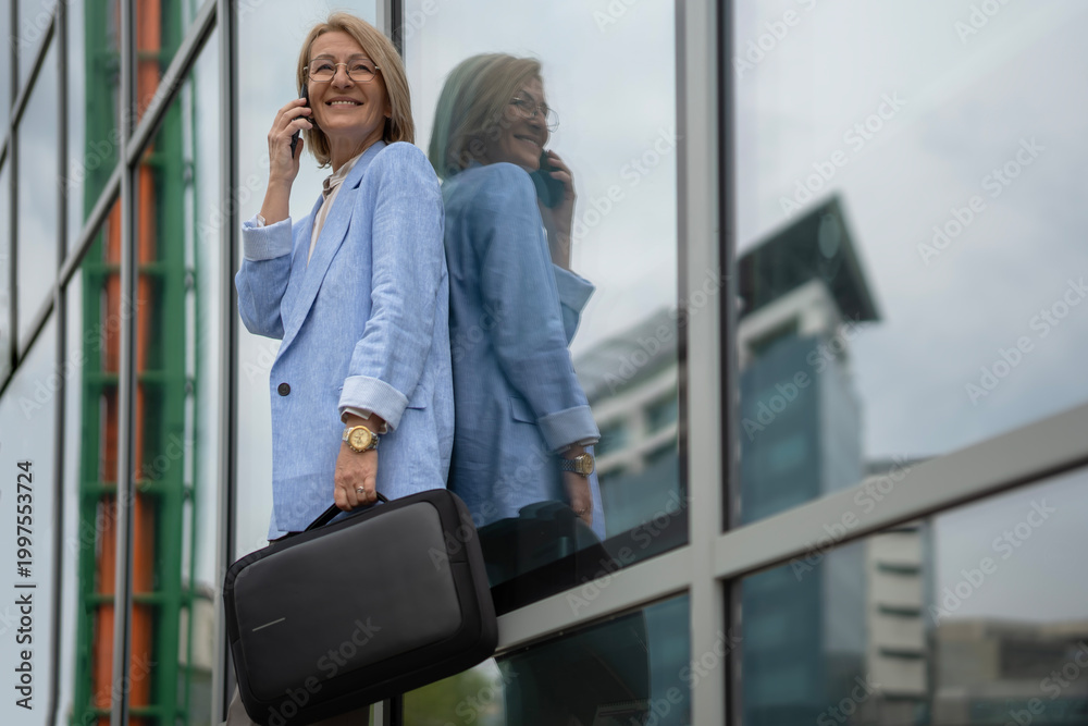 Fototapeta premium Smiling mature businesswoman talking on smartphone outside office