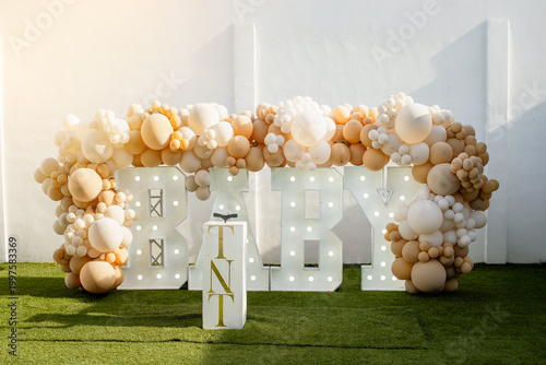 Outdoor setup for a baby gender announcement celebration featuring illuminated letters and balloon arch
