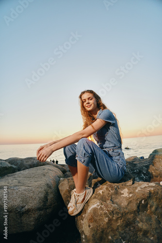lifestyle woman in modern boho-western denim street style sitting on rocks by the sea during warm sunset with film color tones and relaxed vibe