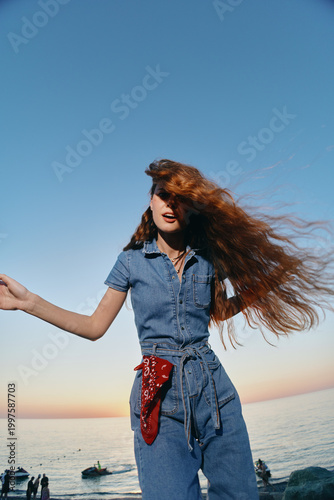 young woman with long red hair wearing denim jumpsuit and red bandana dancing joyfully near the sea at sunset with clear blue sky