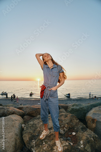 lifestyle woman in modern boho-western denim street style poses confidently on rocky beach during sunset, film color capturing relaxed summer vibe and casual fashion.