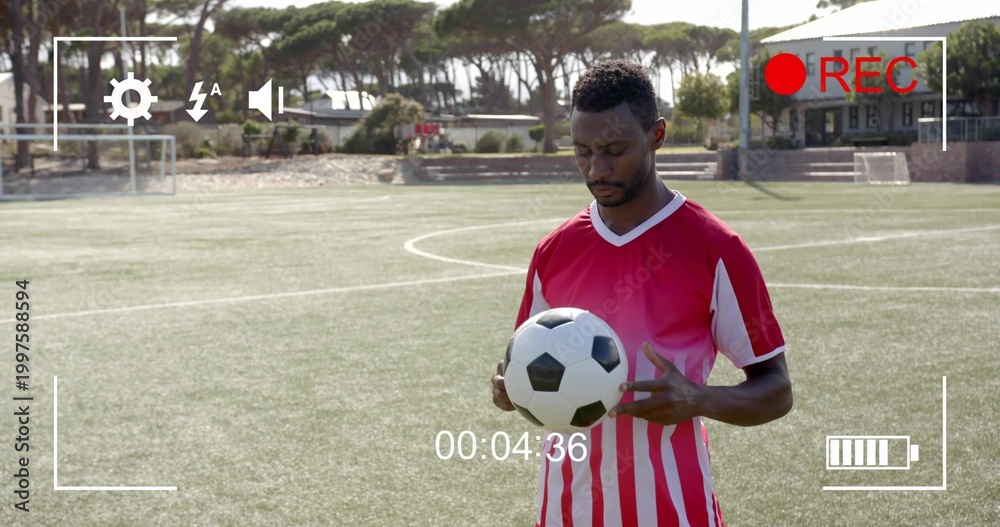 Fototapeta premium Standing man wearing red-and-white kit, holding black-white ball on turf, showing REC 00:04:36