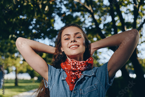 lifestyle woman wearing modern boho-western denim and red bandana in street style with film color effect, smiling confidently under green trees outdoors