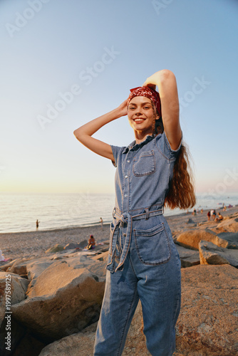 Lifestyle woman in modern boho-western denim street style poses with a red bandana by the beach at sunset in warm film color tones, expressing freedom and joy.