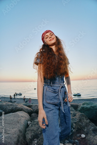 smiling woman with red bandana wearing denim dress standing on rocky beach at sunset with calm sea and boats in background