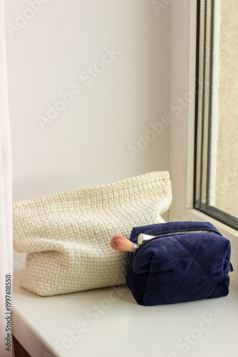 A windowsill scene with a cream woven cosmetic bag, a dark blue quilted makeup pouch with a brush.