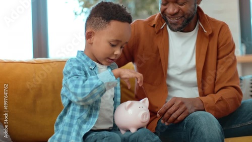 A man with dark skin interacts with a young boy as they save money in a piggy bank. They are sitting on a sofa in a cozy living room, engaging in a positive financial activity.
