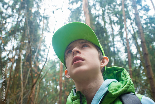 Teenage boy in green cap looking up at tall trees in the forest