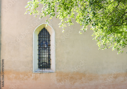 View of a pointed arch window with a metal grate on a textured stone wall partially shaded by green tree leaves Italy.