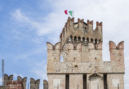 View of Scaligero Castle featuring the medieval stone tower with swallow-tail merlons and the Italian flag waving against a blue sky in Italy.