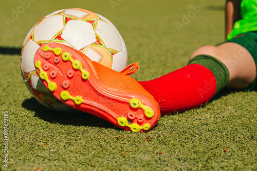 Female football player in green jersey and red socks rests foot on soccer ball on artificial turf field, showcasing bright cleats and vibrant colors in a sports setting