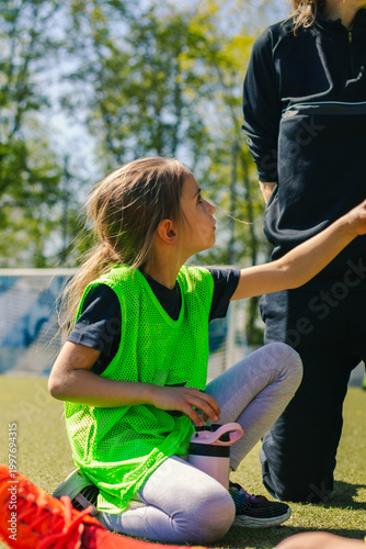 Young girl in bright green training vest kneeling on soccer field while interacting with coach, surrounded by trees and goalposts in the background
