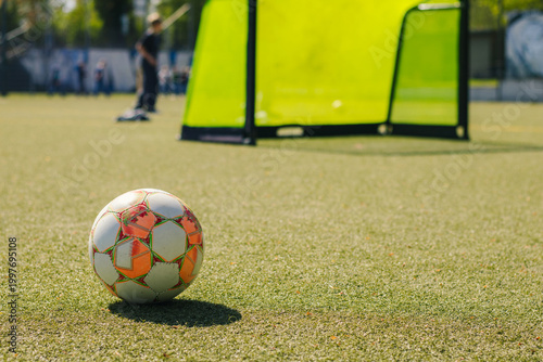 Football on artificial turf with goal net in background, players practicing in distance under clear blue sky during sunny day on sports field