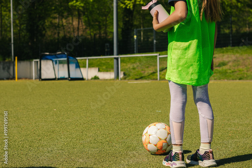 Young girl in green sports jersey stands on a football field, holding a smartphone, with a soccer ball and goalpost visible in the background under bright sunlight