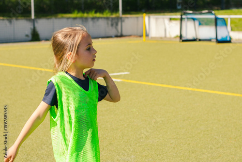 Young girl wearing a green training vest stands on a football field, with goalposts and artificial turf visible in the background under bright sunlight