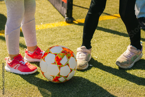 Girls' football scene featuring two pairs of feet in athletic shoes positioned around a colorful soccer ball on a grassy field with a goalpost in the background
