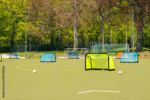 Girls' football training field with multiple small goals and soccer balls arranged on artificial turf under bright sunlight in a green park setting