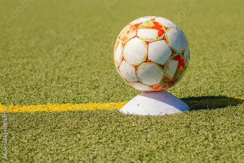 Soccer ball resting on a white cone marker positioned on a green artificial turf field with a yellow line visible in the background