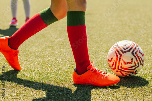 Young female football player in bright orange cleats and red socks dribbles a patterned soccer ball on a green artificial turf field under sunlight