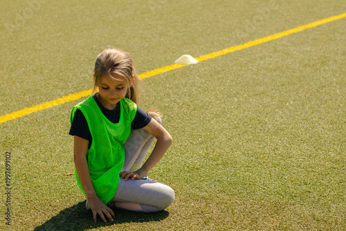 Young girl in green training vest kneeling on artificial turf field, yellow line marking visible, with cones in the background, focused on football practice