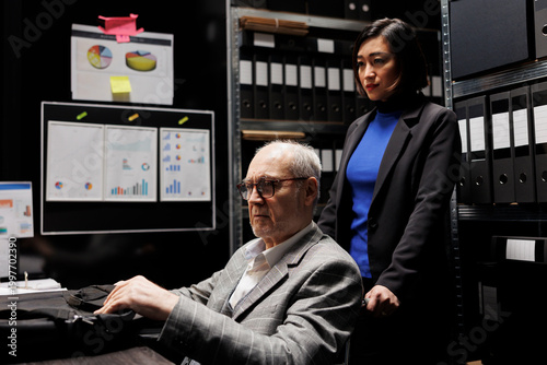 Accountant businessman with disability in wheelchair unpacking laptop before starting shiftwork. Senior executive and asian coworker in archival depository filled with cabinet, folders and flowcharts