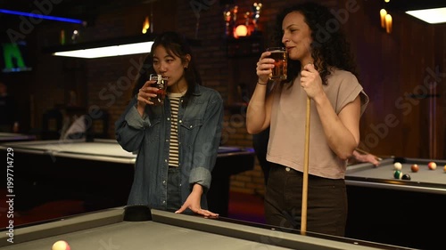 Two women drinking beer and talking while playing pool together in a bar with friends at night