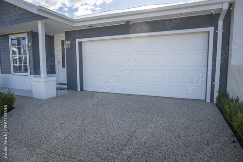 A neat double garage with a white panel lift door and exposed aggregate concrete driveway on a contemporary single-storey Australian home. Modern Australian residential design and curb appeal