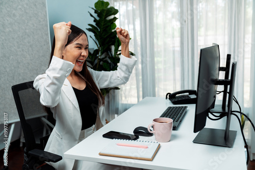 Фотография Successful young Asian businesswoman earning highest profit with fist up in exchange stock market on dynamic business graph on pc screens at modern office, analyzing financial technology