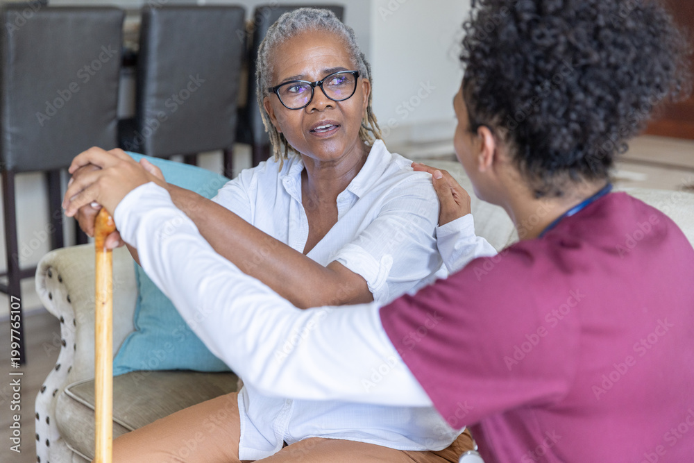Fototapeta premium Senior African American woman with caregiver in maroon scrubs sitting in living room holding cane