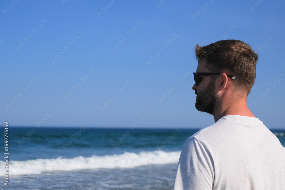 Fototapeta premium Side view of a man in sunglasses and a light T-shirt standing by the ocean with waves under a clear blue sky