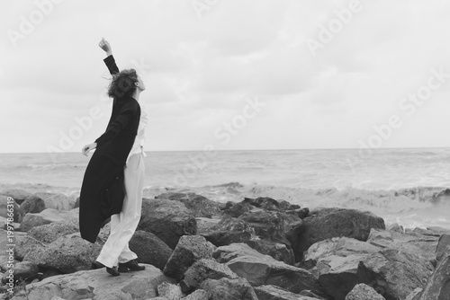 Black and white image of a couple dancing on a rocky shore, wind in coats, embrace mid motion, waves crashing nearby, moody seaside scene conveying freedom, movement and shared escape