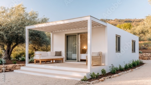A modern, minimalist white house with a shaded porch, set in a serene, sunlit landscape featuring trees and shrubs.