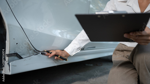 Fotografie Close-up of auto damage inspector examining scratched car body panel with pen and clipboard for insurance claim or repair estimate in a garage