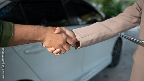 Close-up handshake between two people next to a white car, suggesting agreement during vehicle sale or service transaction, outdoor daylight, shallow depth of field.