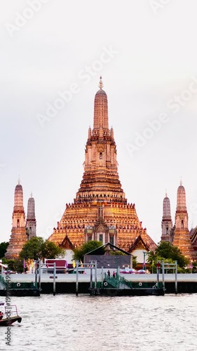 Wat Arun Ratchawararam in Bangkok, Thailand, with a bright golden orange sunset as the backdrop. The reflection of the temple on the water below creates a peaceful and elegant atmosphere.