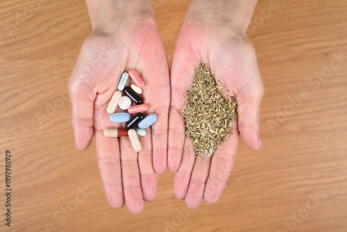 Woman holding capsules and tablets in one hand and heap of dry tea in the other hand over the wooden table. Top view. What to choose: Medicines or herbal treatment?