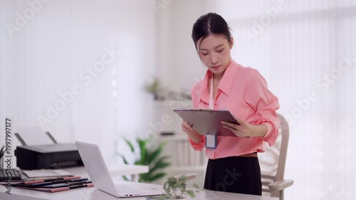 Asian businesswoman reviewing documents standing at office desk using laptop computer multitask professional entrepreneur working at modern workplace checking
