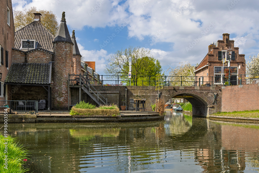 Fototapeta premium Ijsselstein, Netherlands. A picturesque view of an ancient brick building and a small arched stone bridge reflected in the calm waters of a town canal.