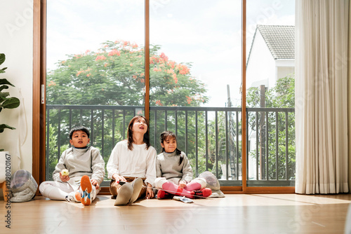 Foto A woman and two kids relax on a sunlit living room floor by large sliding doors, gazing up and smiling, with trees and a balcony outside