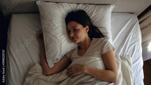 Static overhead shot of woman turning and hugging pillow while sleeping in bed inside cozy bedroom with natural morning light