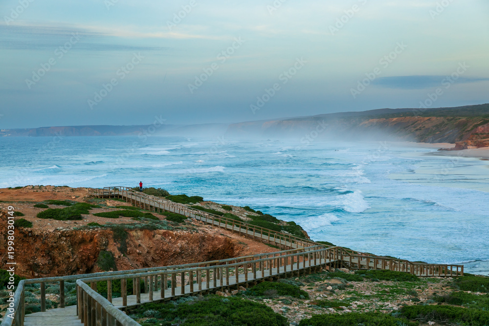 Obraz premium Lone Tourist on Coastal Boardwalk Above Rough Ocean in Algarve