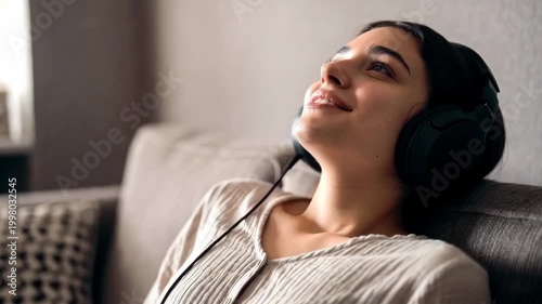 Slow motion static pan of young woman listening and smiling with headphones on sofa in living room daytime