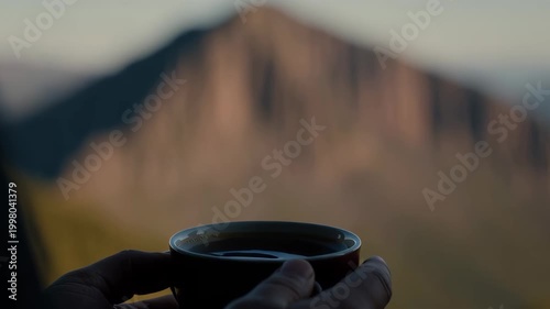 Handheld pull focus shot of hands holding coffee cup as focus shifts from distant mountain ridge to cup on mountain overlook at golden hour