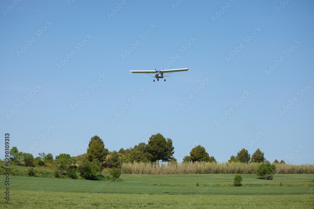 Fototapeta premium Small propeller airplane flying low above green rural field. Light aircraft in clear blue sky over peaceful countryside landscape.