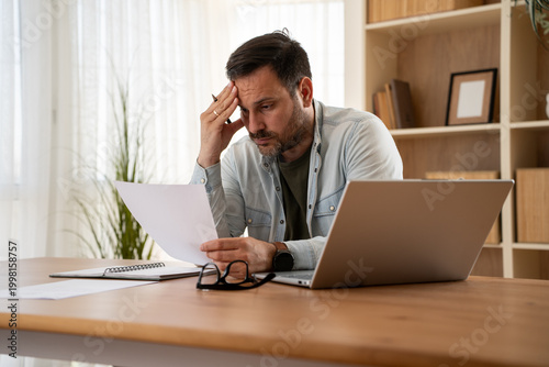 Photos Overwhelmed man handling documents and admin work on laptop at home office