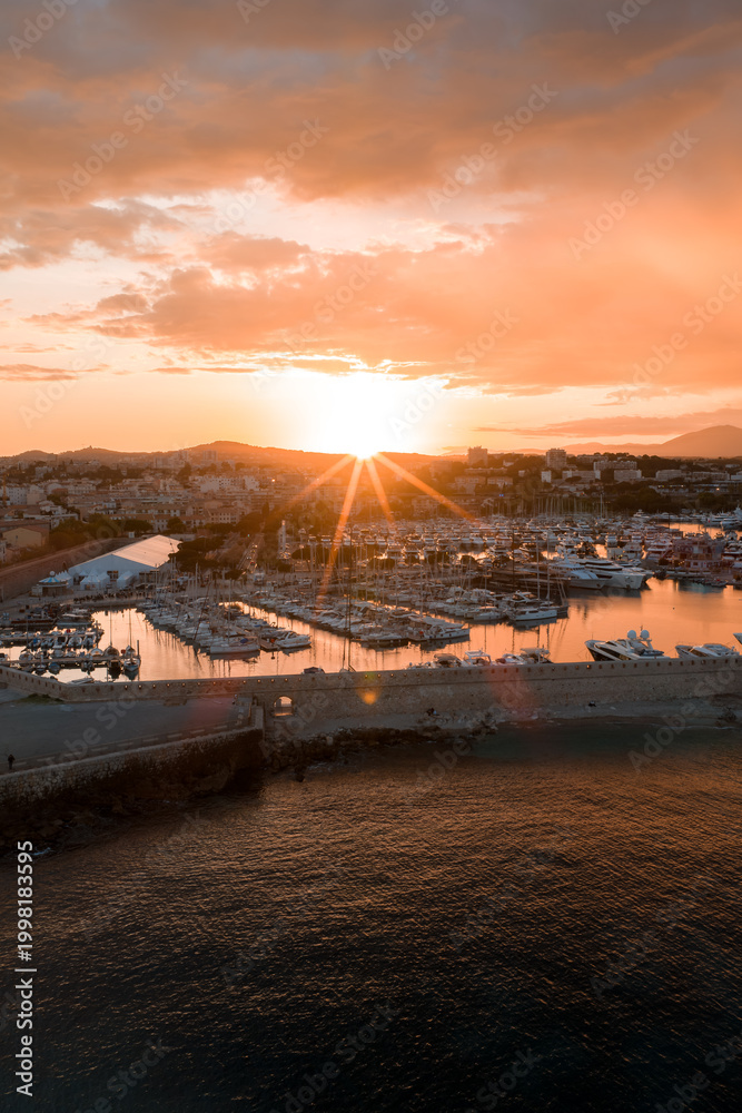 Fototapeta premium Aerial view of Antibes, France at sunset shows the marina with yachts, the compact old town, sandy beaches, and a curved breakwater under soft golden light.