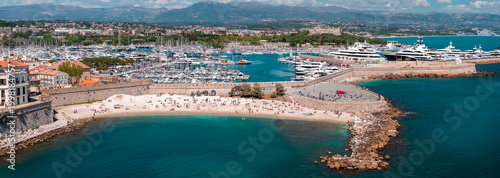 Foto Aerial midday view of Antibes, France, with Plage de la Gravette, stone breakwaters, old town walls, Port Vauban masts, superyachts by IYCA, and faint Alps on horizon