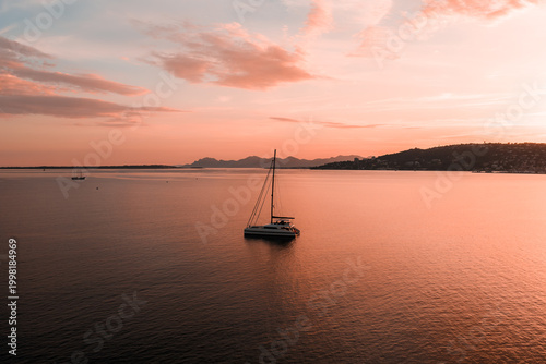 Aerial view of a solitary sailboat drifting on calm rose gold waters near Antibes, France at sunset, pastel sky reflecting across the bay with dusky hills beyond.
