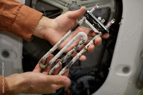 Hands holding burnt washing machine heating element with scale