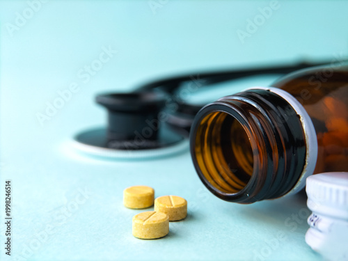 Amber glass bottle filled with round, yellow pills, partially open on a light blue surface. A stethoscope is slightly blurred in the background, suggesting a medical setting. 
